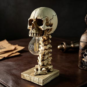 Könnte beinhalten: Dekorative Totenkopflampe mit Glühbirne. Die Lampe wird von einer wirbelsäulenförmigen Struktur und einem Holzsockel getragen. Der Schädel ist cremefarben mit Rissen, die Glühbirne leuchtet warm.