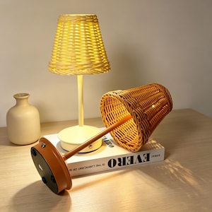 May include: Two wicker lamps with a warm glow. One lamp is upright, the other is tilted, revealing its base. The lampshades are woven, and the bases are round. A small vase is in the background.