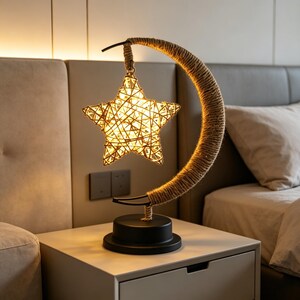 May include: A decorative table lamp featuring a woven star and a crescent moon design. The star is illuminated, and the moon is wrapped in rope. The lamp has a black base and is placed on a white nightstand.