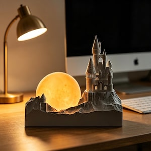 May include: A decorative night light featuring a castle silhouette with a glowing moon. The lamp is gray and has a detailed castle design with multiple turrets. The moon is a warm yellow color, and the lamp sits on a wooden surface.