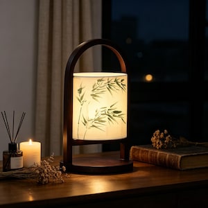 May include: A wooden table lamp with a cylindrical shade featuring a bamboo leaf design. The lamp has a dark brown wooden frame and base. A lit candle and a book are also on the table.