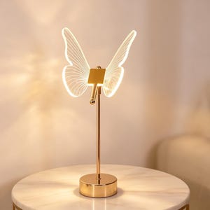 May include: A decorative table lamp featuring a butterfly design. The lamp has a gold-colored base and stem, with a butterfly-shaped light fixture. The butterfly wings are outlined with white lines, and the lamp is illuminated. The lamp is on a white marble table.
