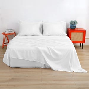 Puede incluir: Una cama con un juego de sábanas blancas y dos almohadas blancas a juego. A la izquierda, un pequeño taburete de madera naranja sostiene una pila de libros. A la derecha, una mesita de noche naranja brillante con frente de rejilla y una maceta azul.