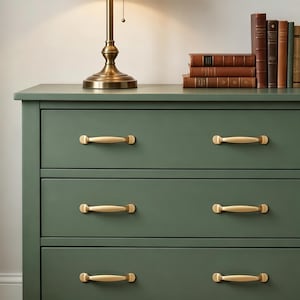 Peut inclure: Une commode vert sauge dotée de trois tiroirs horizontaux, chacun équipé de deux poignées en métal doré brossé. Une lampe de table en laiton et une pile de six livres reliés aux dos marron et bleu sont posées sur le dessus du meuble.