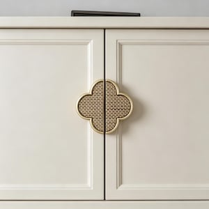 May include: A pair of off-white cabinet doors featuring a central clover-shaped handle. The handle is divided into two symmetrical halves, each framed in gold metal and inlaid with a woven tan rattan texture, creating a decorative accent.
