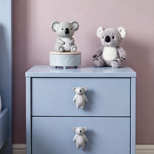 Puede incluir: Una mesita de noche azul claro de dos cajones con dos tiradores en forma de koala. Encima hay una caja de música cilíndrica azul claro con una figura de koala y un peluche de koala gris y blanco con ojos y nariz de color negro.