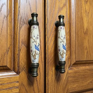 Könnte beinhalten: Zwei Schrankgriffe mit cremefarbenen Keramikgriffen, die jeweils einen blauen Vogel auf einem blühenden Zweig zeigen. Die Metallbeschläge haben eine Bronzefinish. Die Griffe sind an einem Holzschrank montiert.