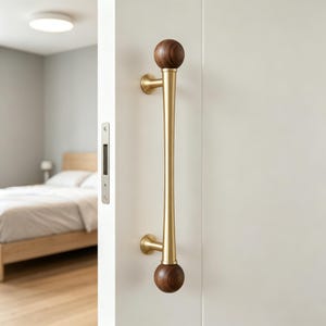 May include: A brass door pull with a wooden ball at each end. The pull is mounted on a white door, with a bedroom visible in the background. The wooden balls are dark brown, and the brass has a polished finish.