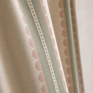 May include: A textured fabric curtain featuring vertical stripes in sage green and cream. The design includes a central band with a small geometric pattern flanked by rows of soft pink scalloped circles on a cream background, creating a layered look.