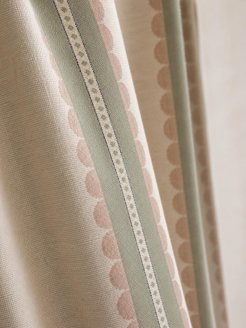 May include: A textured fabric curtain featuring vertical stripes in sage green and cream. The design includes a central band with a small geometric pattern flanked by rows of soft pink scalloped circles on a cream background, creating a layered look.