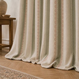 May include: A floor-length curtain featuring vertical stripes in cream, sage green, and soft pink. The fabric displays a repeating pattern of scalloped edges and thin geometric lines, creating a textured appearance with soft, cascading folds.