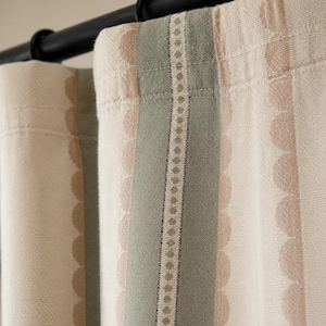 May include: A curtain panel hanging from a black rod with rings, featuring a cream-colored fabric with vertical stripes in sage green and soft beige. The design includes a central decorative band with a repeating diamond pattern and scalloped edges.
