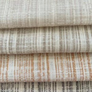 May include: A stack of four folded fabric swatches featuring a textured, woven pattern. The top swatch is cream, the second is light beige, the third is a warm burnt orange, and the bottom swatch is a dark charcoal gray, all with vertical stripes.