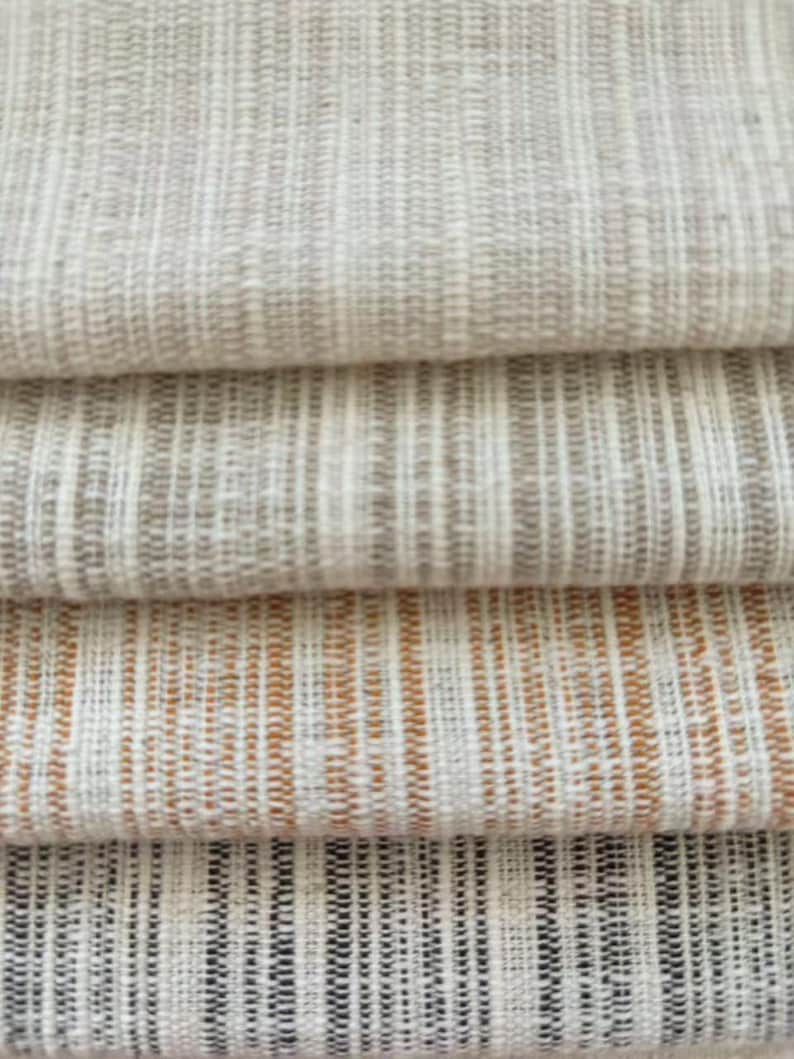 May include: A stack of four folded fabric swatches featuring a textured, woven pattern. The top swatch is cream, the second is light beige, the third is a warm burnt orange, and the bottom swatch is a dark charcoal gray, all with vertical stripes.