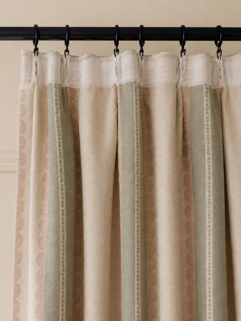 May include: A pleated curtain panel featuring vertical stripes in muted sage green, soft beige, and pale pink. The fabric displays a subtle scalloped pattern within the stripes and is suspended from a black rod by black metal curtain rings.