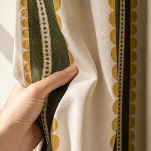 May include: A cream-colored curtain featuring vertical stripes in dark green and mustard yellow. The fabric displays a repeating scalloped edge pattern along the stripes, complemented by a thin, delicate line of small, dark diamond-shaped motifs.