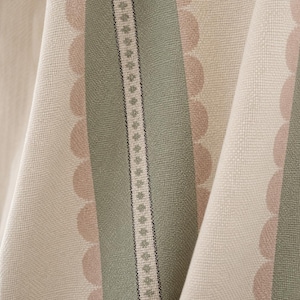 May include: A woven fabric featuring vertical stripes in sage green and cream, accented by rows of soft pink scalloped edges and a central decorative band containing a repeating geometric diamond pattern in green and black on a cream background.