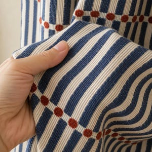 Peut inclure: Un tissu texturé présentant un motif de rayures verticales bleues et crème, entrecoupées de rangées horizontales de petits points en relief de couleur rouille. Le matériau a un aspect côtelé et tricoté avec un motif géométrique répétitif.