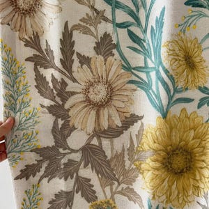 May include: A fabric curtain featuring a botanical print with large yellow and beige daisy-like flowers, dark brown foliage, and teal green stems on an off-white background. The textile has a visible woven texture throughout the floral pattern.
