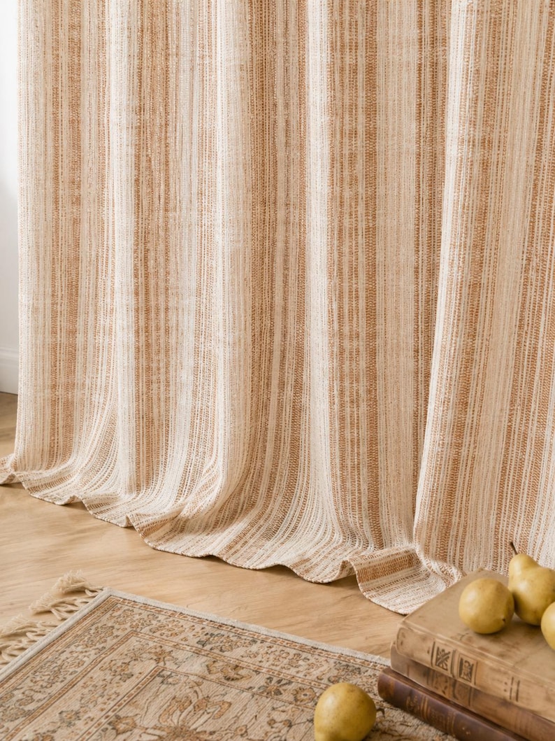 May include: A floor-length curtain featuring vertical stripes in shades of cream and tan. The fabric has a textured, woven appearance with subtle variations in color density throughout the vertical pattern, creating a soft, draped effect.