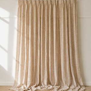 May include: A floor-length curtain with a vertical striped pattern in shades of beige and cream. The curtain features a pleated top header and is suspended from a black metal rod with rings. The fabric has a textured, woven appearance.