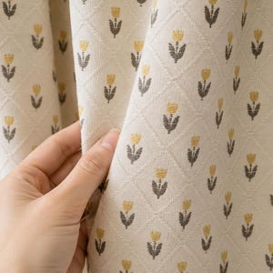 May include: A cream-colored fabric curtain featuring a repeating diamond grid pattern. Each diamond contains a small, stylized yellow and dark gray tulip motif. The textile has a visible woven texture and is draped in soft, vertical folds.