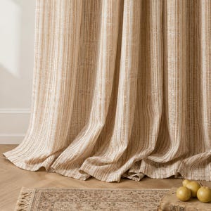 May include: A floor-length curtain featuring a vertical striped pattern in shades of tan, beige, and white. The fabric has a textured, woven appearance with soft folds that drape down to the floor, creating a layered effect in the lower section.