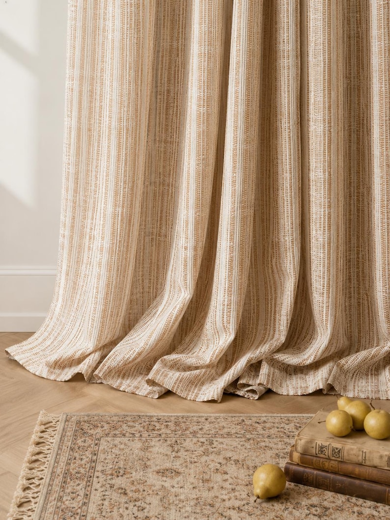 May include: A floor-length curtain featuring a vertical striped pattern in shades of tan, beige, and white. The fabric has a textured, woven appearance with soft folds that drape down to the floor, creating a layered effect in the lower section.