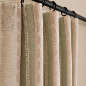 May include: A curtain panel featuring vertical stripes in muted sage green and soft beige, accented by a repeating scalloped pattern in a light pink hue. The fabric is suspended from a black metal rod using circular rings with clips.