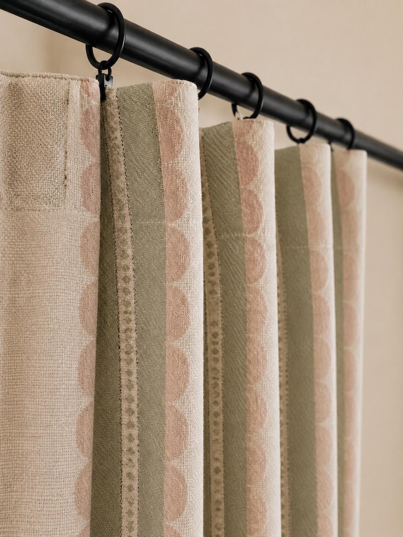 May include: A curtain panel featuring vertical stripes in muted sage green and soft beige, accented by a repeating scalloped pattern in a light pink hue. The fabric is suspended from a black metal rod using circular rings with clips.