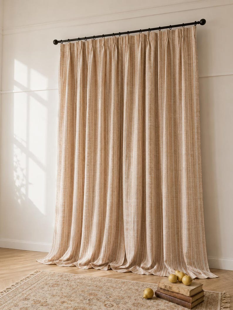 May include: Floor-length curtains featuring a vertical striped pattern in shades of beige and cream. The fabric has a textured appearance and hangs from a black metal curtain rod with circular rings, creating soft, uniform pleats along the length.