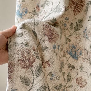 Könnte beinhalten: Ein gewebter Stoff mit cremefarbener Basis und einem sich wiederholenden Blumenmuster. Das Design zeigt detaillierte Blüten und Blätter in gedeckten Tönen von Altrosa, Salbeigrün und Schieferblau, was eine strukturierte Optik erzeugt.
