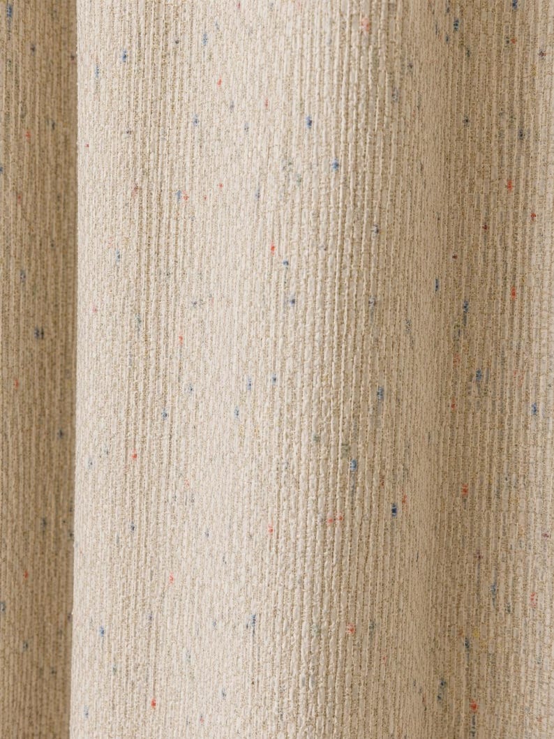 以下が含まれることがあります： 縦のリブ模様と、素材全体に散りばめられた青、赤、緑の小さな斑点が特徴の、質感のあるベージュの布製カーテン。生地は柔らかい縦のひだを描いて垂れ下がっており、織りの深みと触感の良さを際立たせています。