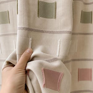 May include: A beige fabric curtain featuring horizontal textured stripes and a repeating pattern of small, square-shaped patches in muted sage green, soft pink, and cream colors, with visible frayed edges on each square patch.