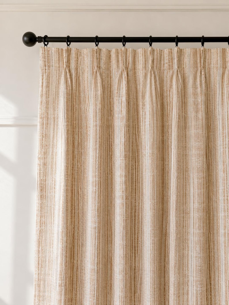 May include: A floor-length curtain panel featuring a vertical striped pattern in shades of tan, beige, and white. The fabric has a textured weave and is suspended from a black metal rod with matching black rings, creating uniform pinch pleats.