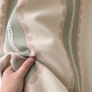 May include: A woven fabric featuring a pattern of vertical sage green and soft pink scalloped stripes. The design includes thin white lines with small dot details running between the colored bands, set against a cream-colored background.