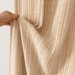May include: A curtain panel featuring a vertical striped pattern in shades of tan and off-white. The fabric has a visible woven texture with alternating thin and thick lines running vertically throughout the length of the material.