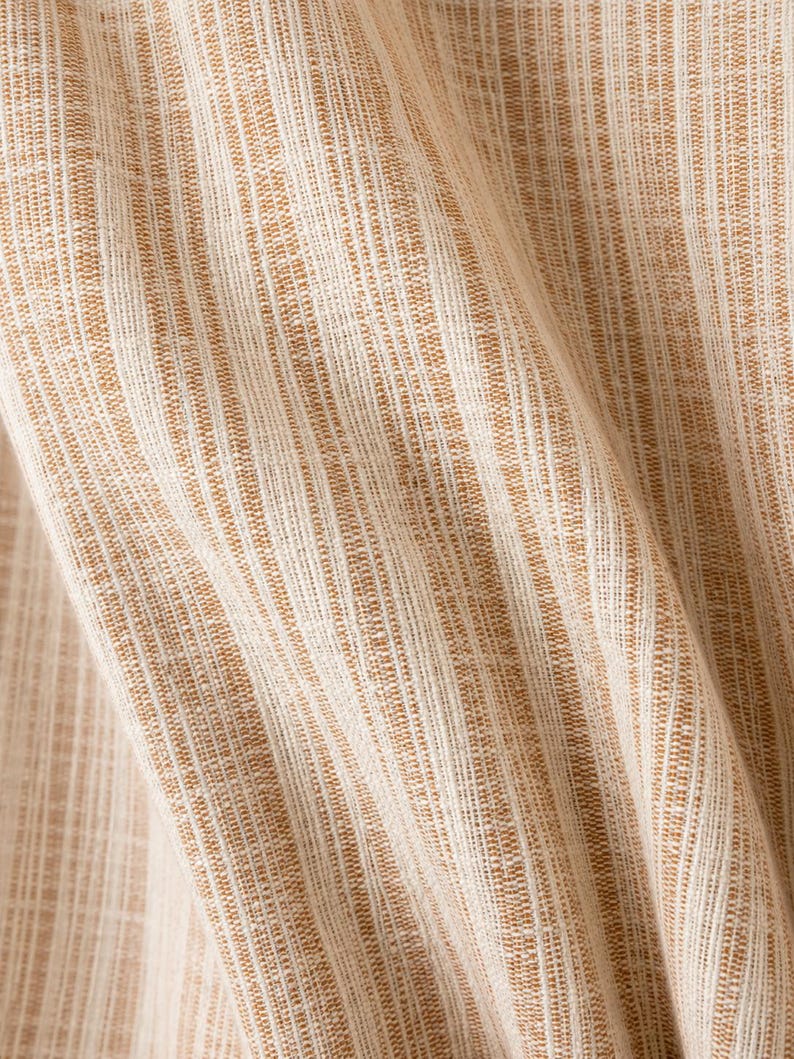 May include: A woven fabric featuring a vertical striped pattern in shades of tan and off-white. The material displays a textured, slubbed appearance with visible fibers running in parallel lines, creating a consistent, repeating linear design throughout.