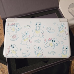 Könnte beinhalten: Eine weiße tragbare Spielkonsole mit einem wiederkehrenden Muster aus blau umrandeten Cartoon-Krokodilen mit gelben Brustakzenten und roten Rückenstacheln, durchsetzt mit kleinen Fischen, Seerosenblättern und Kleesymbolen auf der Oberseite.