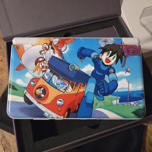 Puede incluir: Consola de juegos portátil con una vibrante ilustración de un personaje de dibujos animados con un traje azul corriendo sobre un fondo verde y azul. El diseño incluye un vehículo rojo y naranja y otros personajes.