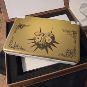 May include: Gold-colored handheld gaming console featuring a detailed Majora's Mask design. The console has white accents and decorative corner elements. The design is a stylized illustration with a dark brown color. The console is inside a wooden box.