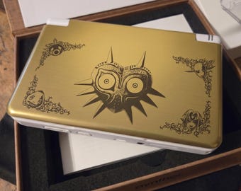 Può includere: Console di gioco portatile dorata con un design di Majora's Mask. La console ha dettagli bianchi ed elementi decorativi angolari. Il design è un'illustrazione stilizzata con colore marrone scuro. La console è all'interno di una scatola di legno.