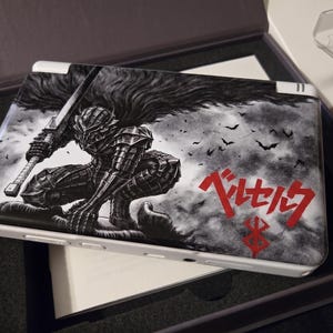May include: A white handheld gaming console with a black and white graphic of a warrior holding a sword, with the word "Berserk" in red Japanese text. The console is in a black box.
