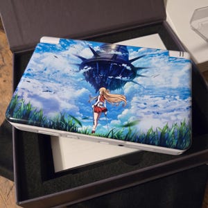 May include: A white handheld gaming console with a custom cover featuring anime artwork. The cover depicts a female character with long blonde hair, a red skirt, and a sword, set against a backdrop of a cloudy sky and a large, dark structure.