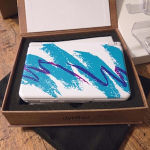 May include: A white handheld gaming console featuring a teal and purple abstract brushstroke pattern on the top cover. The device is housed in a brown box with the text "aynthor" printed on the front edge, resting in a black foam insert.