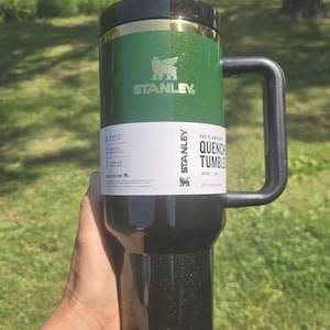K&ouml;nnte beinhalten: Ein gr&uuml;ner und schwarzer Stanley Quencher Becher mit Griff. Der Becher hat einen schwarzen Deckel und ein wei&szlig;es Etikett mit dem Stanley-Logo und Produktinformationen. Der Becher wird in einer Hand gehalten.