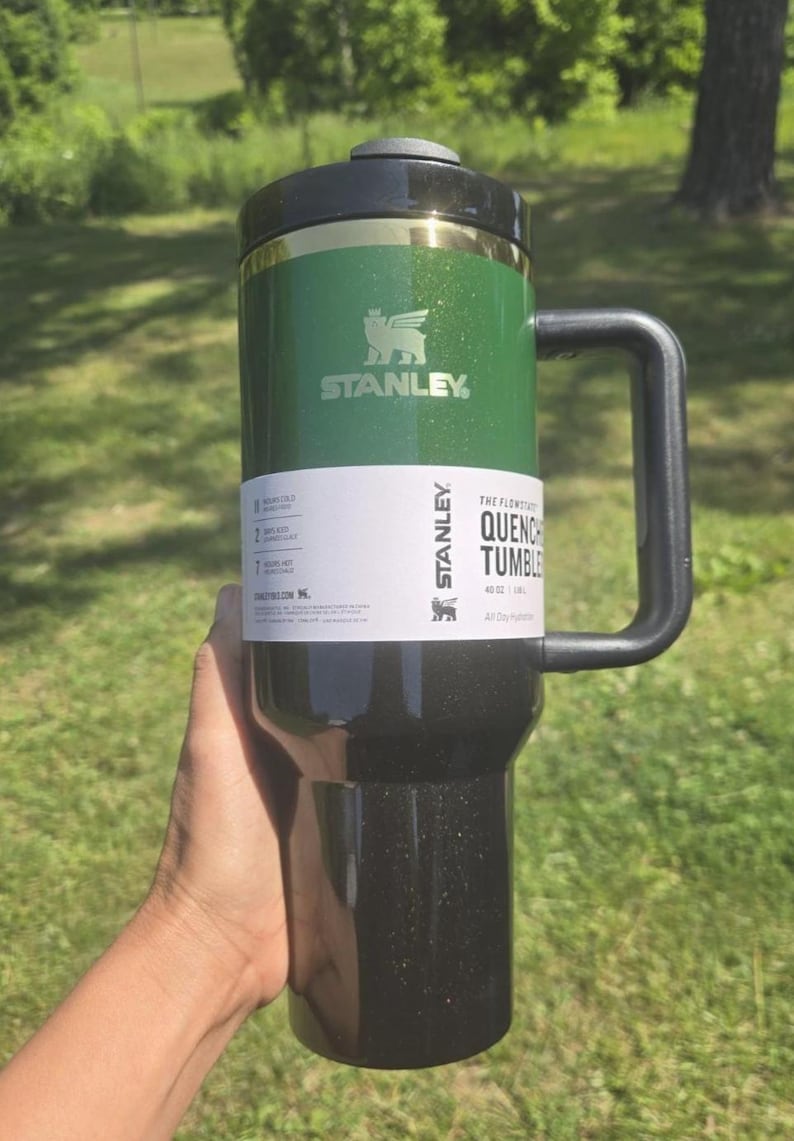 K&ouml;nnte beinhalten: Ein gr&uuml;ner und schwarzer Stanley Quencher Becher mit Griff. Der Becher hat einen schwarzen Deckel und ein wei&szlig;es Etikett mit dem Stanley-Logo und Produktinformationen. Der Becher wird in einer Hand gehalten.