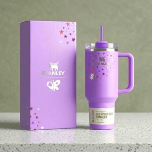 以下が含まれることがあります： ラベンダー色のスタンレーQuencher H2.0タンブラーと、お揃いの箱。タンブラーにはハンドル、ストロー、蓋が付いています。タンブラーと箱の両方に星と蝶のアクセントがあります。箱とタンブラーにはスタンレーのロゴがあります。