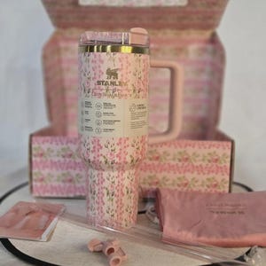 Op de afbeelding: Een roze Stanley-beker met bloemenpatroon, handvat, rietje en accessoires, gepresenteerd met een bijpassende doos en etui. De beker heeft de tekst "STANLEY" en "LOVESHACKFANCY".