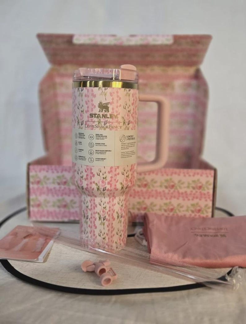 Op de afbeelding: Een roze Stanley-beker met bloemenpatroon, handvat, rietje en accessoires, gepresenteerd met een bijpassende doos en etui. De beker heeft de tekst "STANLEY" en "LOVESHACKFANCY".